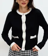 Cardigan de punto color blanco con detalles en negro en cuello, puños y tapeta frontal. Presenta cierre frontal con botones perlados y dos bolsillos delanteros falsos con solapa bicolor.