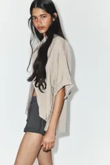 Camisa de corte fluido confeccionada en mezcla de modal, con cuello solapa, manga corta y bolsillo de plastrón en el delantero. Presenta cierre frontal con botones ocultos por tapeta y bajo con aberturas laterales.