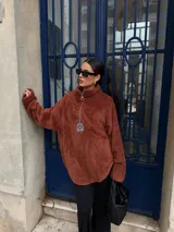 Buzo de corderito color marrón, con cuello alto y cierre frontal parcial. Presenta un diseño holgado con mangas largas y un parche decorativo en el frente.