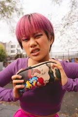 Estuche pequeño con forma de media luna, estampado camuflado en tonos tierra y verde militar, con un diseño gráfico abstracto y colorido superpuesto. Posee cierre superior con tirador de cremallera negro.