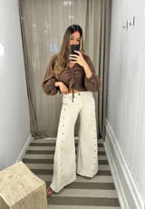 Pantalón de pierna ancha color crudo, confeccionado en lino, con diseño de botones metálicos a lo largo de los laterales y ruedo desflecado.