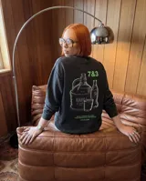 Buzo de cuello redondo en color negro con efecto lavado, de corte regular y ajuste cómodo. Presenta un estampado gráfico en la espalda con motivos de bebidas y texto alusivo, en tonos verde neón y violeta.