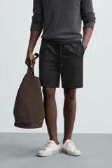 Bermuda negra de corte recto, con cintura elástica ajustable mediante cordón, pliegues frontales y bolsillos de vivo en la espalda. El bajo tiene un dobladillo vuelto.