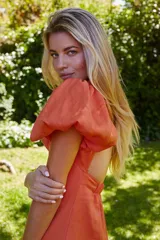 Vestido color naranja con escote redondo, largo asimétrico y espalda descubierta con cierre de botón. Tiene mangas cortas abullonadas con elástico.