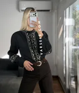 Camisa de manga larga color blanco con bordados geométricos en negro a lo largo de la botonadura frontal y en los puños. Presenta cuello mao y cierre frontal con botones.