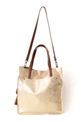 Bolso tipo tote de cuero con acabado metalizado en tono dorado. Cuenta con doble asa de mano y una correa larga ajustable y extraíble en color marrón. Presenta un bolsillo frontal externo y detalle de borlas laterales.