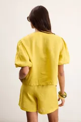 Blusa de lino color amarillo, con cuello redondo, mangas cortas abullonadas y cierre frontal con botones.