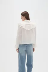 Camisa de algodón blanca con cuello camisero, cierre frontal de botones y volados bordados en el frente y hombros. Presenta mangas largas con puños acampanados y detalles bordados en el ruedo.