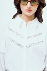 Camisa blanca de manga larga abullonada con puños elásticos, cuello camisero clásico y cierre frontal de botones. Presenta detalles de alforzas y encaje geométrico en la pechera.
