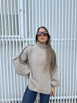 Buzo oversize de punto color beige, con cuello alto, mangas largas abullonadas y detalles de costuras decorativas bordadas a mano en color negro a lo largo de los hombros y mangas.