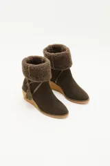 Bota de caña media color verde oliva, confeccionada en cuero con detalle de solapa superior en lana rizada. Presenta un cordón decorativo con borlas en el mismo tono y una suela de plataforma tipo cuña en color beige.
