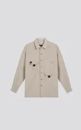 Camisa de manga larga color beige, estilo técnico con calce oversize. Presenta cuello camisero, cierre frontal de botones y múltiples bolsillos de parche con solapa en el frente, algunos con tiradores negros.