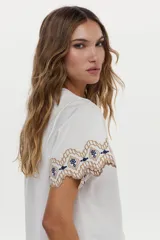 Blusa blanca de jersey con mangas cortas amplias de tela plana, decoradas con bordados geométricos en tonos marrón y azul marino.