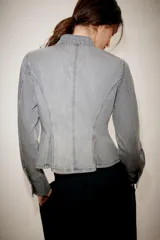Chaqueta de denim gris con cuello subido y manga larga. Presenta un diseño entallado con bajo en evasé y un llamativo bordado frontal de alamares en color negro. Cierre frontal con corchetes ocultos.