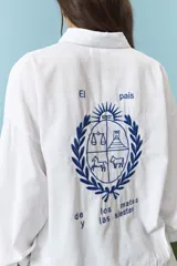 Camisa blanca de corte oversize con cuello camisero y mangas largas. Presenta un bordado en la espalda con el diseño de un escudo nacional estilizado en color azul y la frase 'El país de los mates y las siestas'.