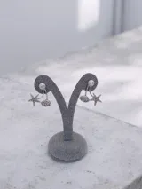 Par de caravanas colgantes de metal plateado con diseño temático marino. Presentan una perla superior y dos dijes colgantes: una estrella de mar y una concha marina, ambos adornados con strass.