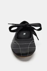 Zapato plano tipo bailarina de estilo deportivo, color negro, con pespuntes blancos contrastantes en el cuerpo y puntera. Presenta un escote redondo y un lazo decorativo en el empeine. La suela es fina y de color marrón claro.