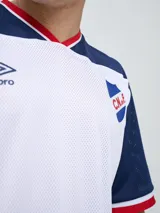 Remera de fútbol Umbro del Club Nacional de Football, de diseño tricolor con cuerpo azul marino, mangas y canesú rojos, y cuello en V con detalles en blanco y azul. Incluye el logo de Umbro en el pecho derecho y el escudo del club en el izquierdo.