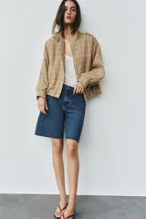 Chaqueta bomber de color beige con diseño a cuadros blancos, cuello subido, manga larga con trabillas en los hombros, bolsillos delanteros y bajo con elástico. Cierre frontal con cremallera y botones a presión.