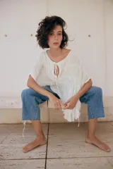 Top blanco de cuello redondo y mangas tipo capa, con textura fruncida, bajo asimétrico, lazos decorativos en el escote y en los laterales, y cierre posterior con botón.