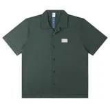 Camisa de manga corta color verde oscuro con cuello cubano y cierre frontal de botones negros. Presenta un pequeño parche rectangular bordado en el pecho izquierdo con texto y gráficos.
