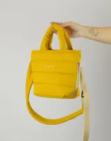 Bolsito tipo tote acolchado de color amarillo, con diseño de líneas horizontales y logo de la marca estampado en el frente. Cuenta con dos asas superiores acolchadas y una correa ajustable de cinta en color beige.