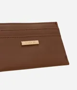 Portatarjetas pequeño de color marrón, confeccionado en poliuretano (PU) con textura lisa. Presenta tres ranuras visibles para tarjetas y una placa metálica dorada con el logo de la marca en el frente.
