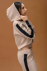 Conjunto de dos piezas en color beige con detalles en negro, compuesto por un buzo con capucha y cierre frontal, y un pantalón de corte recto y holgado. Ambas prendas presentan franjas laterales en contraste.