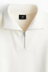 Buzo de tejido pesado color crema, con interior cepillado suave. Presenta cuello elevado, medio cierre metálico y hombros caídos. Los puños y el bajo son de punto acanalado.