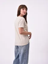 Remera de manga corta color beige claro con cuello redondo. Presenta un estampado gráfico central con un plato de pasta rodeado por un borde azul y blanco, sobre un fondo de franjas verticales rojas y blancas. El estampado incluye texto en cursiva en tonos turquesa y rojo.