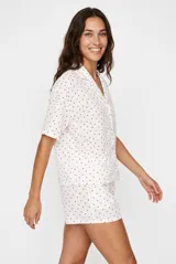 Pijama de dos piezas para mujer, compuesto por una camisa de manga corta con solapas y botones frontales, y un short a juego, ambos confeccionados en tejido blanco con diseño a rayas verticales sutiles y estampado de pequeños corazones rojos.