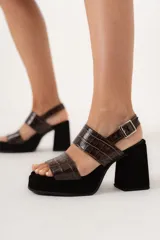 Sandalias de tacón alto con plataforma, color negro y marrón con estampado de cocodrilo. Tienen dos tiras anchas sobre el empeine y una tira con hebilla alrededor del tobillo. El tacón es ancho y cuadrado.