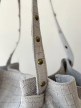 Cartera tipo shopper de cuero gris con textura símil cocodrilo, diseño amplio, correas ajustables con tachas y cierre superior.