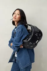 Cartera de hombro color negro, confeccionada en cuero sintético, con diseño de apliques en forma de flor decorados con tachas metálicas plateadas.