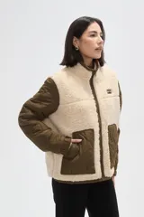 Campera de diseño híbrido que combina paneles de corderito sintético en color crema con mangas y bolsillos de tela técnica acolchada en color verde militar. Presenta cierre frontal completo, cuello alto y un pequeño parche decorativo en el pecho.