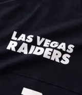 Remera deportiva de corte oversized, color negro, con estampado gráfico en la espalda del equipo Las Vegas Raiders de la NFL. Presenta cuello redondo, mangas cortas con detalles de rayas blancas y logo de la NFL en la parte superior trasera.