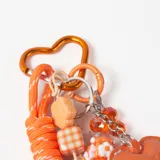 Llavero decorativo en tonos naranja, compuesto por una cuerda trenzada, un mosquetón en forma de corazón y múltiples dijes colgantes que incluyen cuentas de cristal, figuras de flores, un dije de corazón de cuero sintético y una esfera brillante.