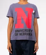 Remera de manga corta, color gris malva, con gran estampado frontal de la letra 'N' en rojo y el texto 'UNIVERSITY OF NEBRASKA' en negro. Presenta un lavado especial y terminaciones desgastadas en cuello, mangas y ruedo.
