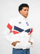 Buzo deportivo de nylon con medio cierre frontal y cuello alto. Presenta un diseño de bloques de color en azul, blanco y bordó, con el escudo del Club Nacional de Football y el logo de Umbro en el pecho.