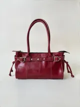 Cartera de cuero color bordó, de estructura rígida y tamaño medio. Presenta una tira decorativa frontal con tachas metálicas y hebillas en los extremos, además de lazos laterales. Cuenta con dos asas superiores para llevar al hombro y cierre principal.