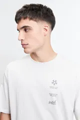 Remera de manga corta, corte oversize, color blanco o hueso, con cuello redondo y estampado gráfico en el pecho que incluye un símbolo de estrella, texto "NORREBRO STUDIOS INTERNATIONAL", "MISTRAL 76 Resilience" y "Distrikt ARCHIVE".