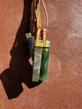 Collar con dije cilíndrico de resina verde con inclusiones blancas, engarzado en metal dorado y colgante de cordón de cuero marrón.