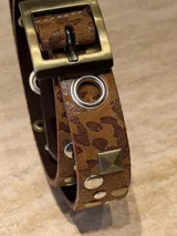 Cinto de cuero con estampado animal print, decorado con tachas metálicas, remaches y ojales plateados. Cuenta con una hebilla metálica cuadrada en tono bronce envejecido.