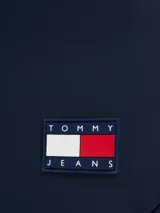Mochila azul marino con acabado liso, compartimento principal con cierre de cremallera, bolsillo frontal vertical con cremallera, dos bolsillos laterales para botellas y correas ajustables. Incluye parche engomado con el logo de Tommy Jeans en la parte delantera.