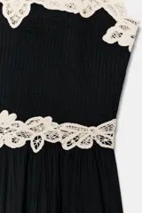 Top negro con escote pico y tirantes finos. Presenta detalles de encaje calado color crema en el escote y a la altura de la cintura, con una caída plisada en la parte inferior.