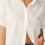 Camisa de manga corta blanca con bordados al tono y diseño calado de puro algodón, con cierre frontal de botones y terminación inferior ondulada.