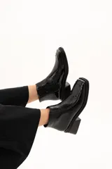Bota corta de cuero negro con acabado brillante, diseño de punta cuadrada y taco bloque. Presenta cierre lateral interno y detalle de ribete en la capellada.