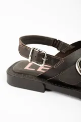 Sandalias de cuero marrón oscuro, con tiras cruzadas en el empeine, hebilla metálica grande decorativa y tira ajustable en el talón con hebilla plateada.