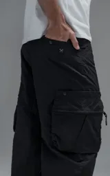 Pantalón cargo oversize de corte relajado, confeccionado en textil técnico resistente de color negro. Presenta bolsillos laterales cargo de gran tamaño con cierres metálicos, bolsillos laterales superiores y bolsillos traseros con broches a presión. Incluye cinturón integrado de cinta de seguridad con hebilla de encastre técnica.
