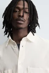 Camisa de algodón con estructura canvas, color rojo lavado, de corte relajado (relaxed fit) y manga corta. Posee cuello solapa, dos bolsillos de plastrón en el pecho y cierre frontal de botonadura. Presenta bordado y estampado de texto a contraste en el delantero.